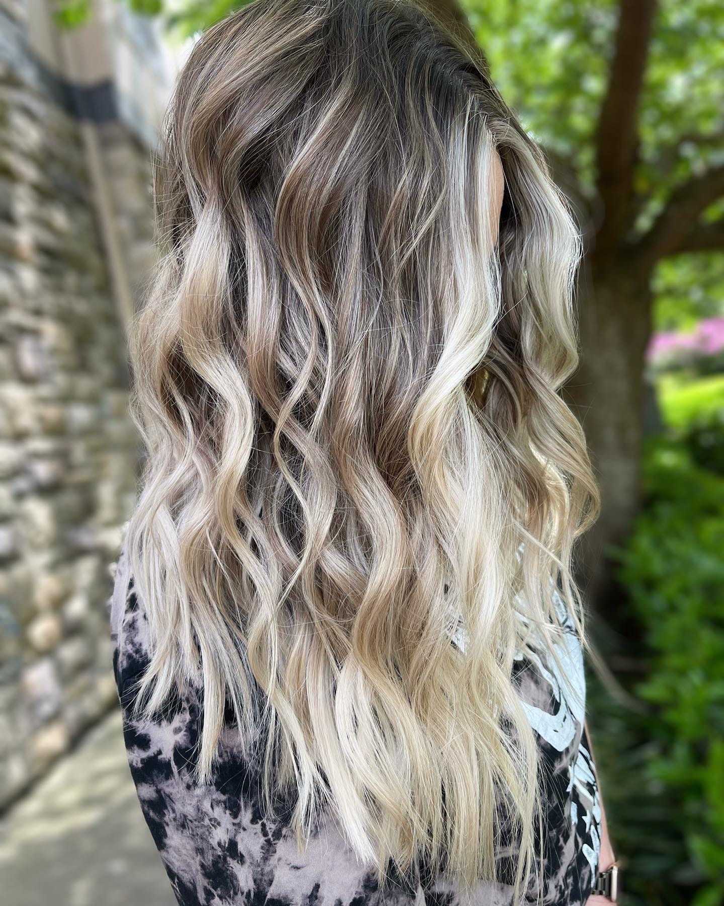 Wavy, blonde hair with dark roots seen outdoors near a stone wall and greenery.