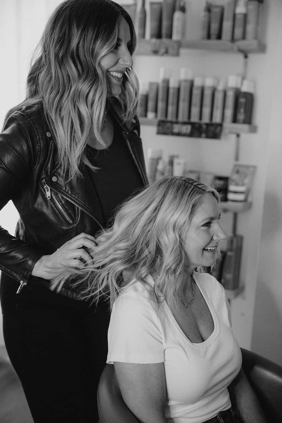 Hairdresser styling a woman's hair in a salon, both smiling and relaxed.